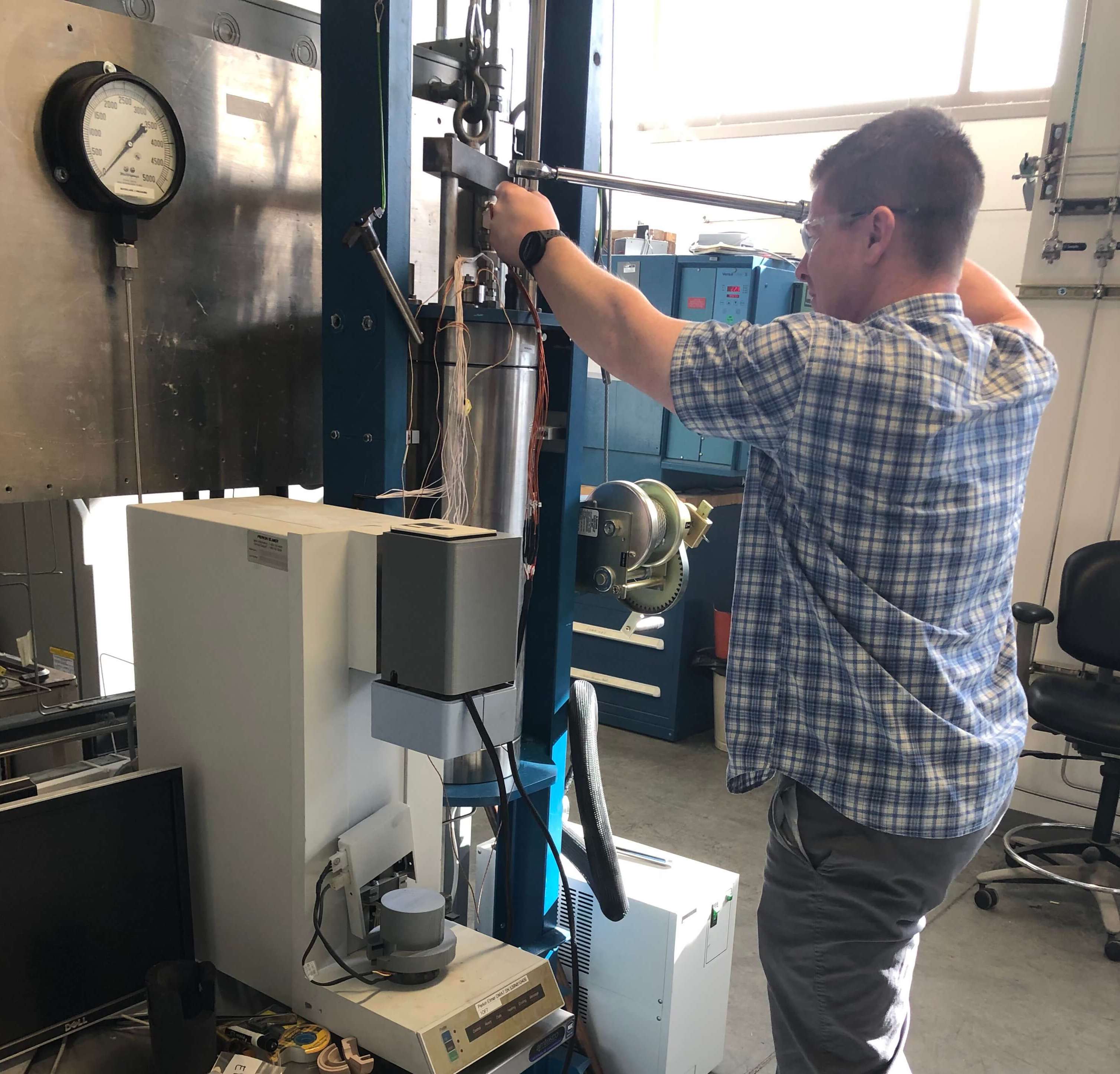 A man wearing a short sleeved checkered shirt, jeans, and safety glasses tightens a bolt with a socket wrench. The bolt is part of a piece of lab equipment which looks like a large metal cylinder suspended between two vertical metal bars